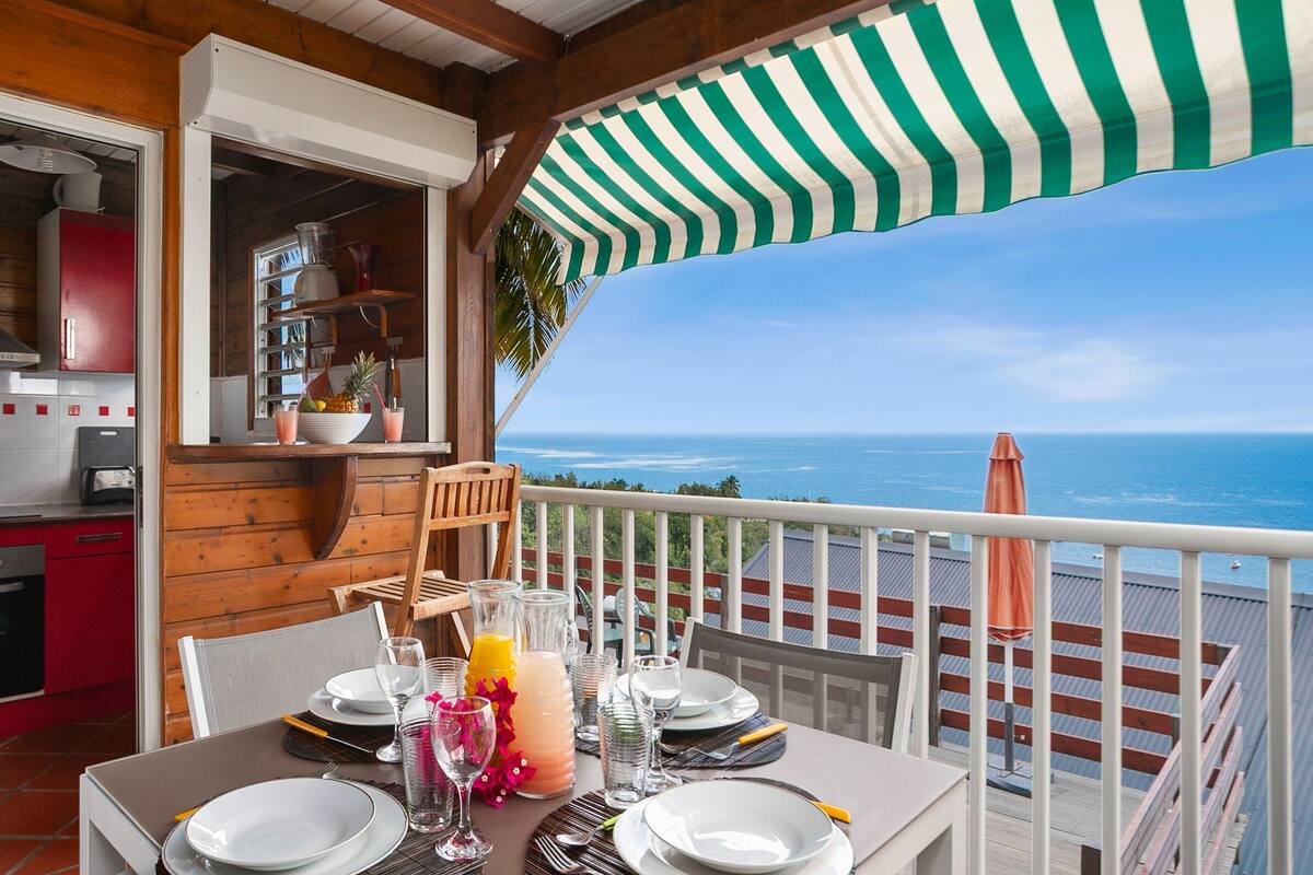 Mesa de comedor para cuatro con vista al océano, una zona de cocina y un toldo a rayas.