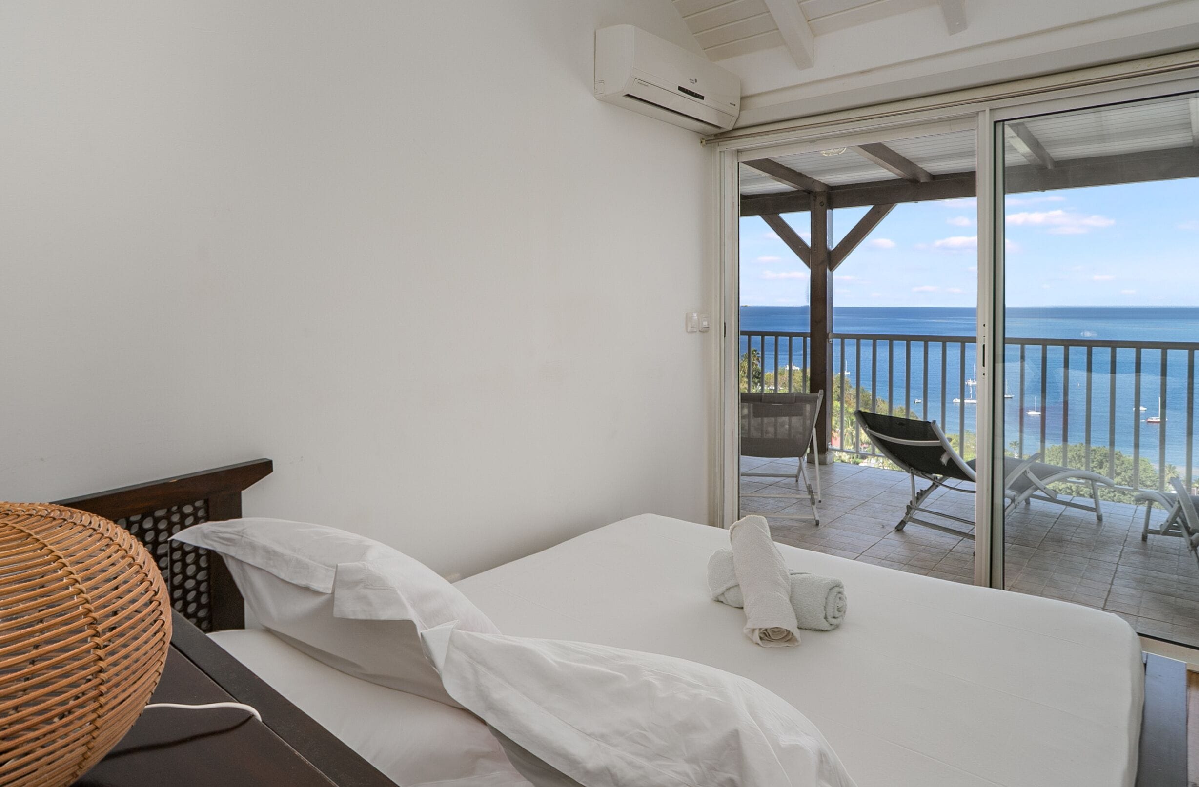 A bedroom with a bed, a wicker chair, and a view of the sea through a sliding glass door.