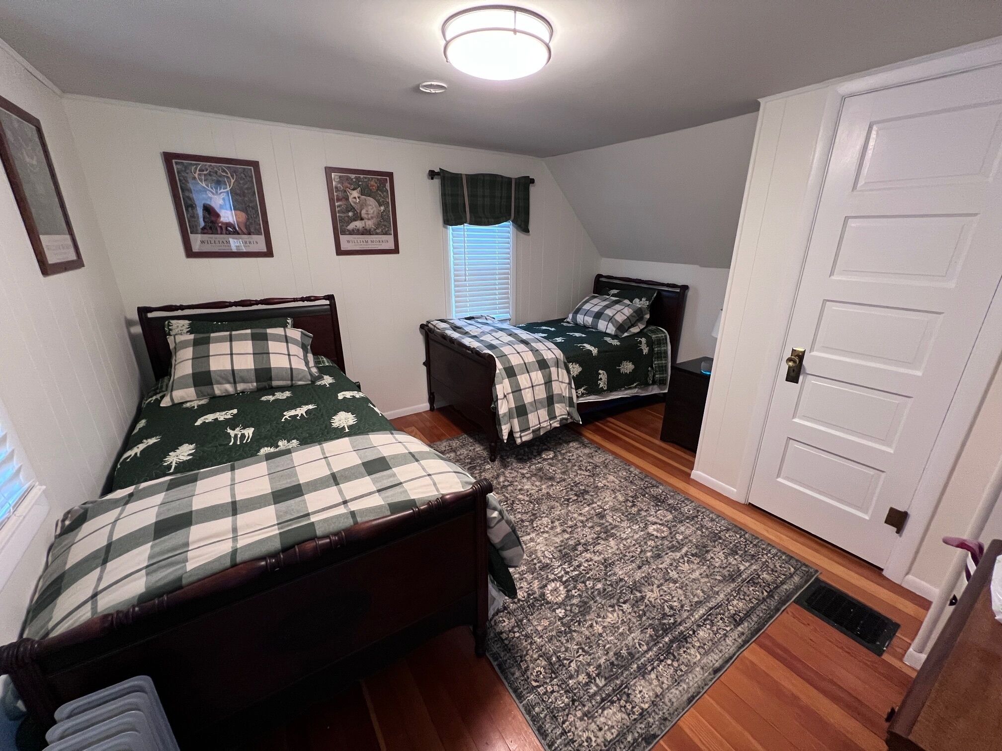 A bedroom with two beds, a window with blinds, a door, and a ceiling light.