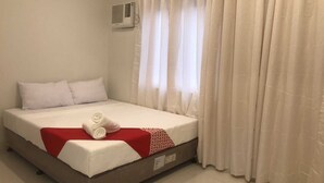 Una cama bien hecha con ropa blanca y un detalle rojo, un aire acondicionado de pared y cortinas blancas traslúcidas.