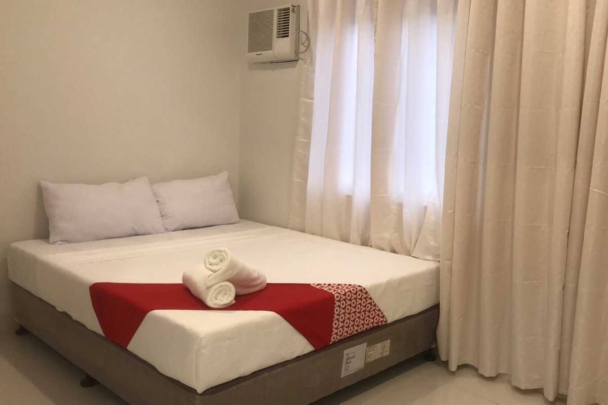 Una cama bien hecha con ropa blanca y un detalle rojo, un aire acondicionado de pared y cortinas blancas traslúcidas.