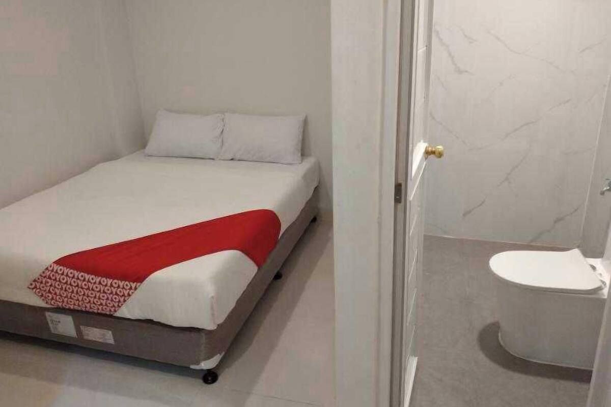 Una cama individual con una colcha roja, un almohadón blanco y un cubrecolchón blanco. Junto a la cama hay un inodoro blanco.