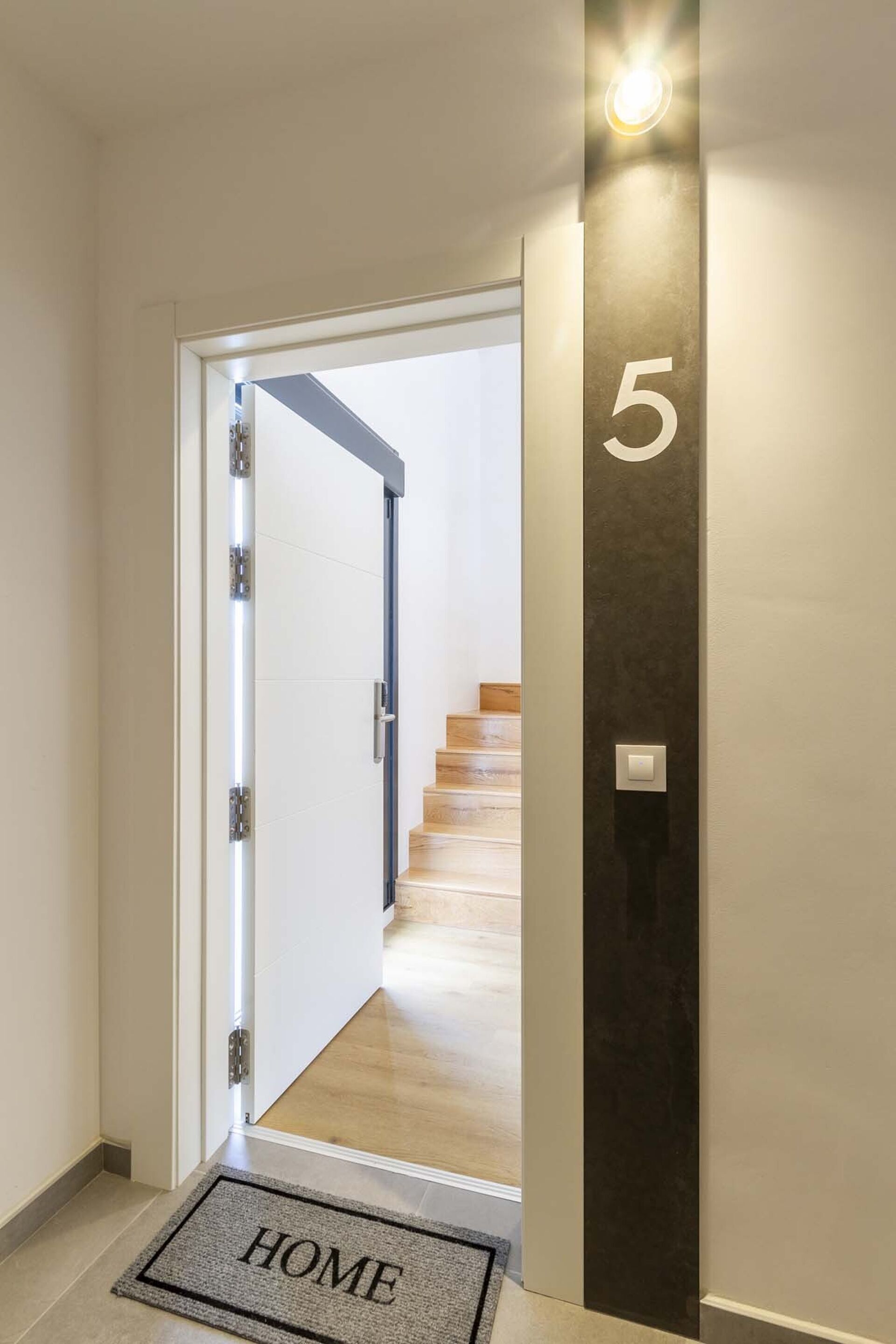 Ein Flur mit einer zur Treppe führenden Tür, einer Fußmatte mit der Aufschrift 'HOME' und einer an der Wand befestigten Nummer 5.