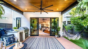 A modern patio with a ceiling fan, outdoor seating, and a view into a well-lit interior space.