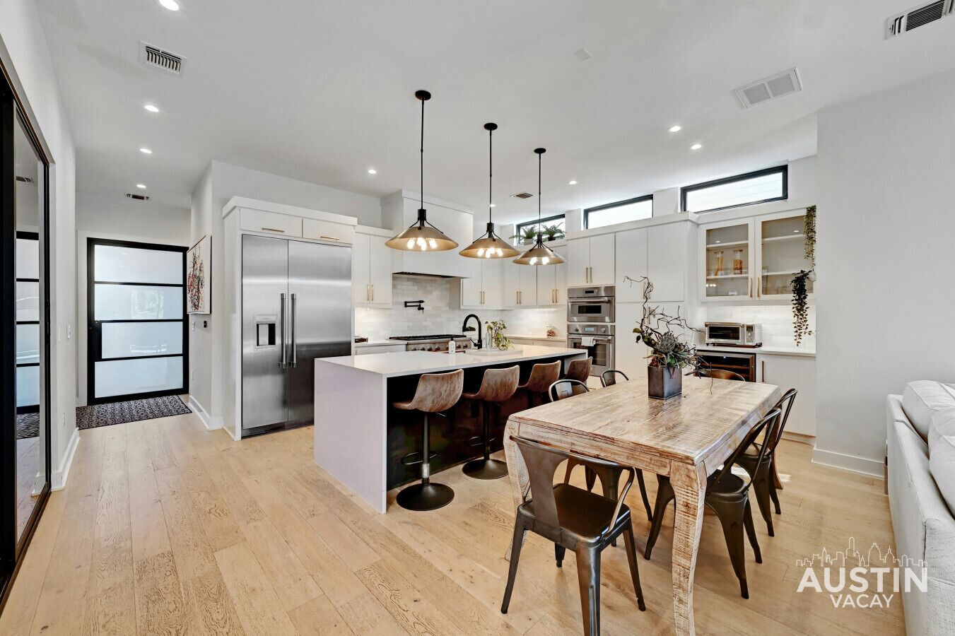 A modern kitchen with a central island, stainless steel appliances, pendant lighting, and a dining area with wooden chairs.