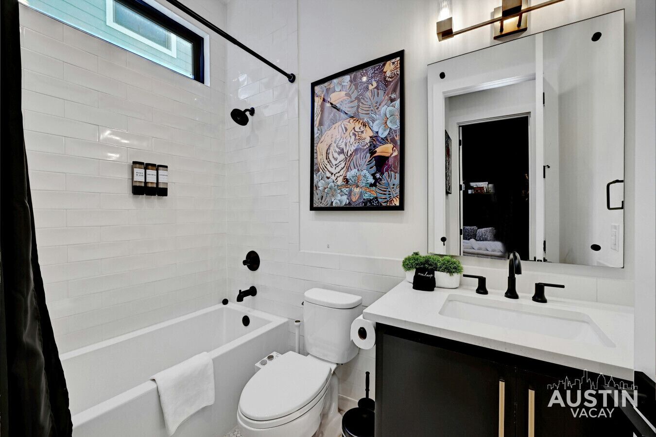 A modern bathroom with a white tiled wall, a bathtub, a toilet, and a sink with a mirror above it. There is a framed artwork on the wall and a small plant on the sink.