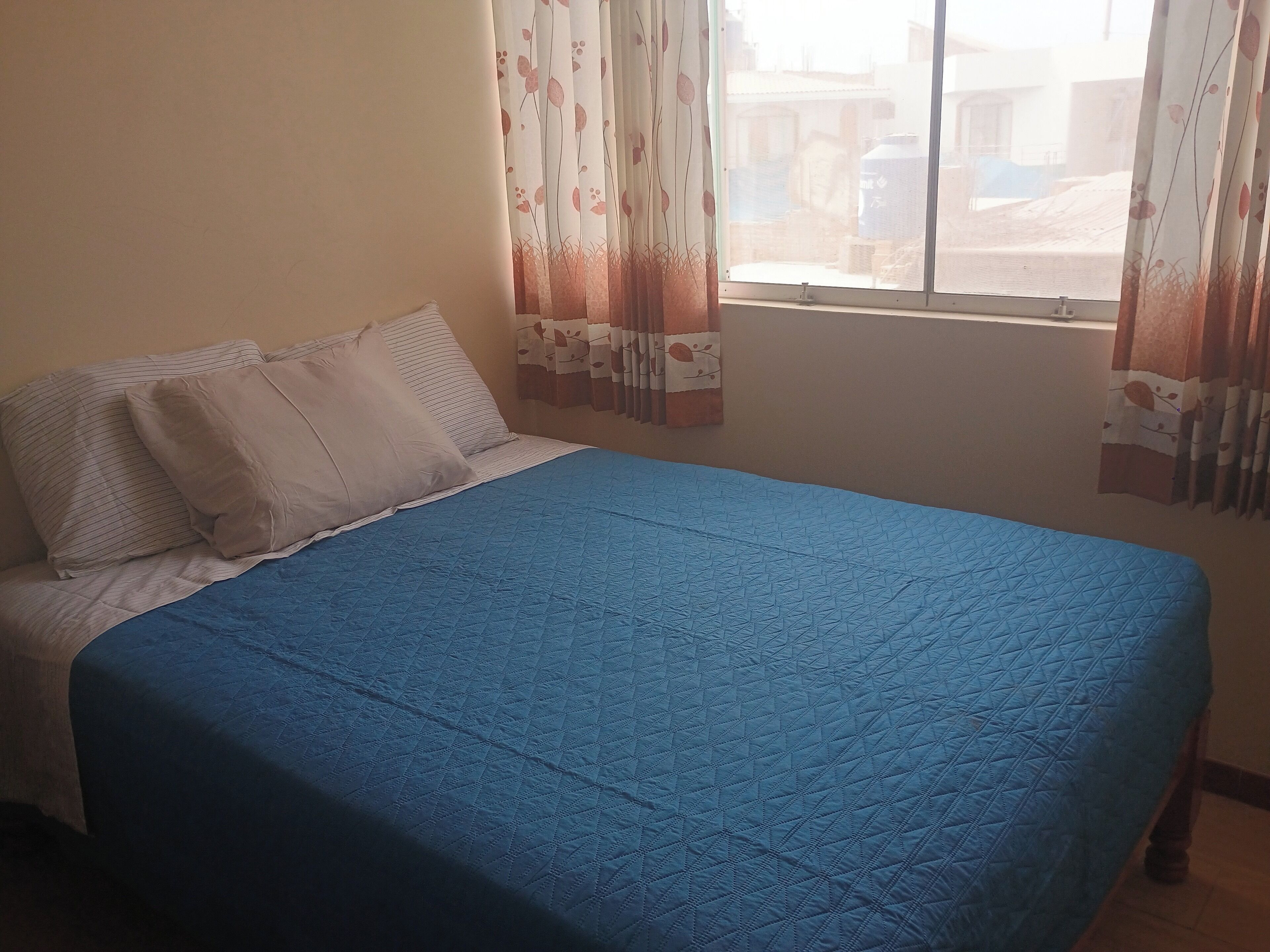 Een bed met een blauwe deken, witte kussens en een raam met bloemen gordijnen.