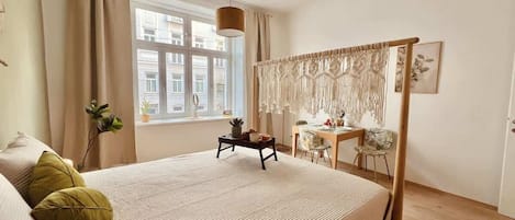 A bedroom with a bed, a desk, a chair, a macrame curtain, and a window with a view of buildings.