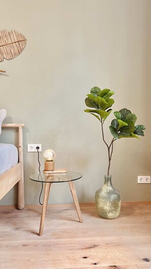 A room with a bed, a small round table with a lamp, and a large green plant in a vase.