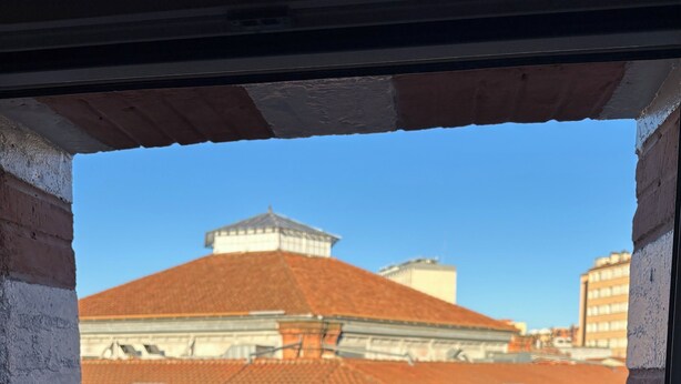Vista a través de una ventana con barandilla de metal, mostrando un edificio de techo con tejas y paredes de piedra.