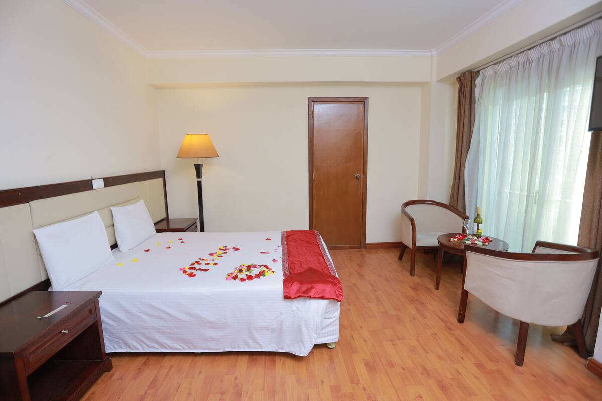 Habitación de hotel con cama, mesita de noche, lámpara, silla y una pequeña mesa con una botella y flores.