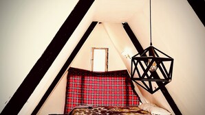 A small, triangular room with a bed, a red plaid pillow, and a geometric hanging light fixture.