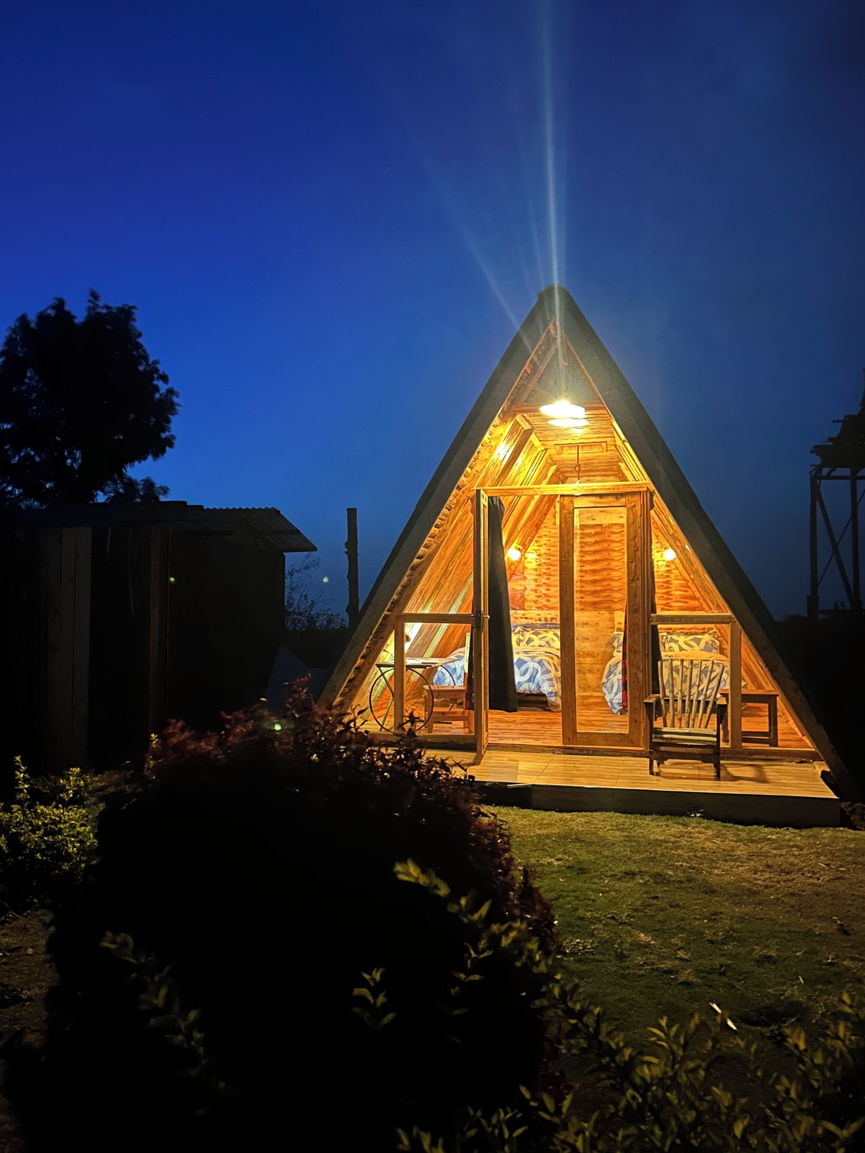 A triangular wooden structure with a bed and seating area, surrounded by a garden.