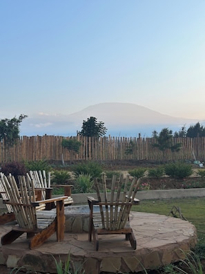A stone patio with two wooden chairs and a table, overlooking a mountain and a wooden fence. - Amboseli Trails Cozy farmhouse with Kilimanjaro Views (Kajiado County)
