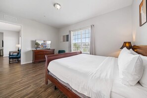 Ein Schlafzimmer mit einem hölzernen Bett, einem Schminktisch, einem Sessel, einem Fernseher und einem Fenster mit Vorhängen.