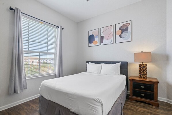 A bedroom with a bed, a nightstand with a lamp, a window with curtains, and wall art.