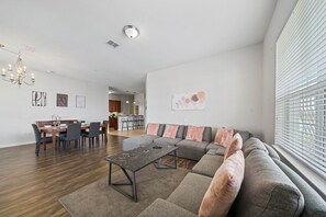 A modern living room with a sectional sofa, a dining table with chairs, a chandelier, and artwork on the walls.