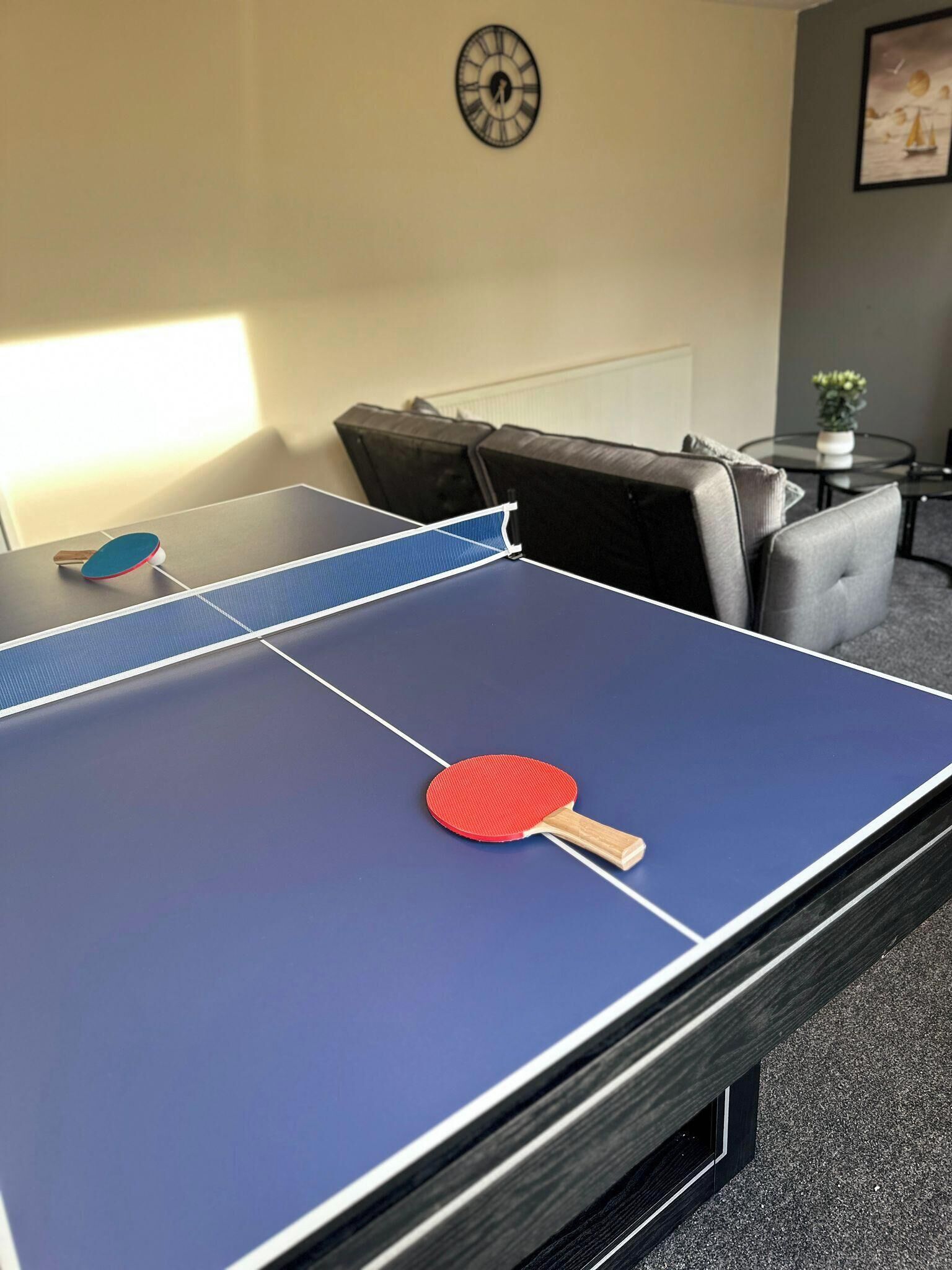 A ping pong table with two paddles, a clock, and a framed picture on the wall.