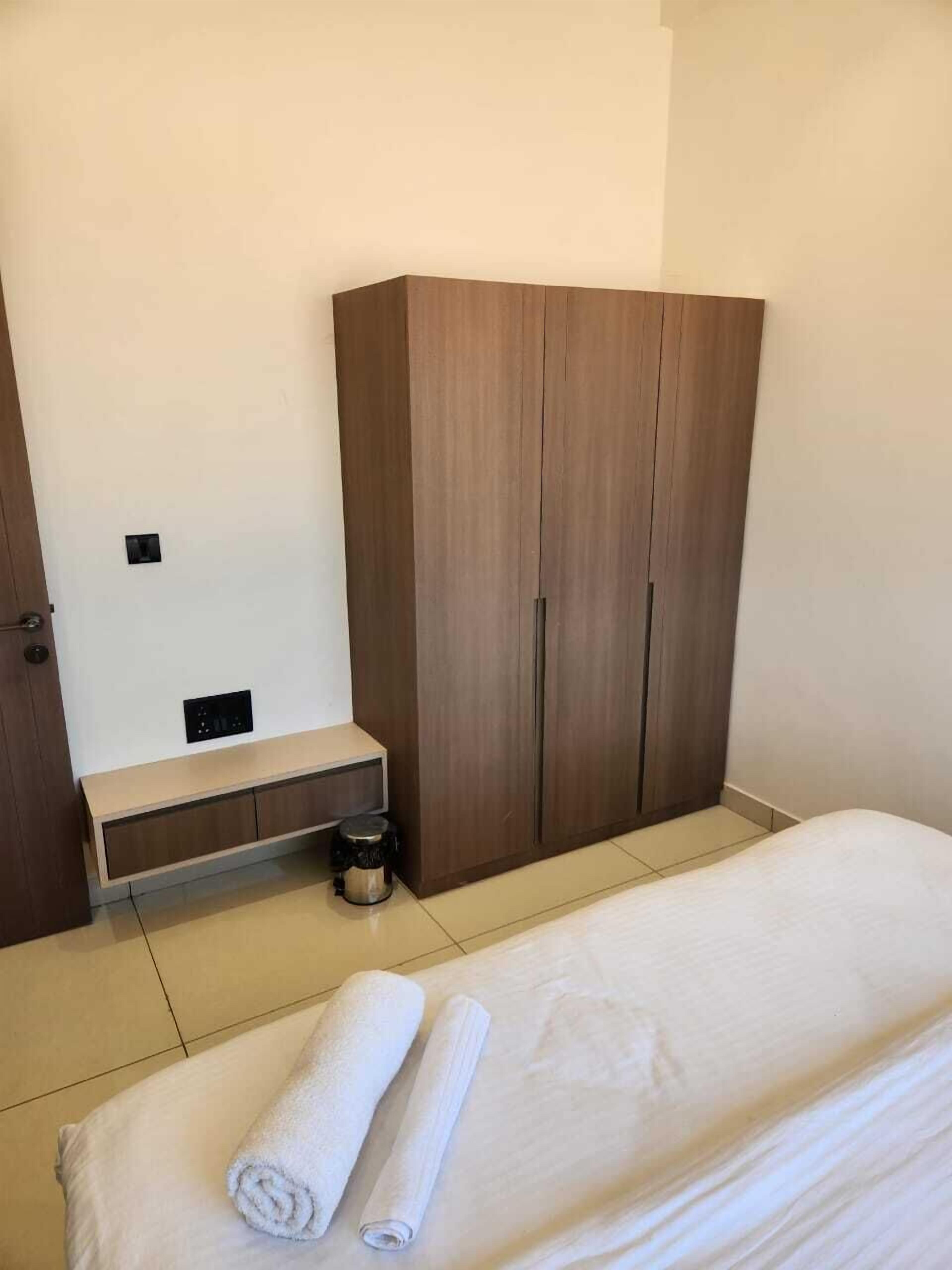Un dormitorio con una cama, dos toallas enrolladas, un armario de madera, un banco y un pequeño cubo de basura.