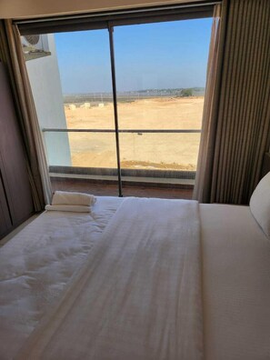 A hotel room with a large window offering a view of a sandy landscape and distant buildings. - Shivam Hotel-Madhavpur (Porbandar)