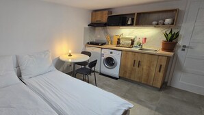 A compact room with a bed, a washing machine, a small table with a candle, and a kitchenette with cabinets and a microwave. - Vu's Home - Apartment Garden - 30m2 (Hlavní město Praha)