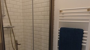 Une douche fermée par des parois de verre, avec des murs recouverts de carreaux blancs, un porte-serviettes sur lequel est posée une serviette bleue, et un radiateur blanc.