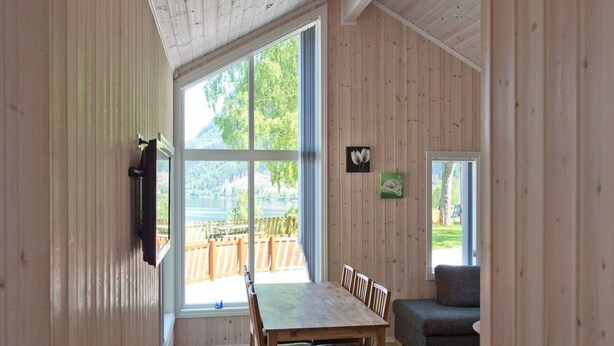 Et træpanelt interiør med spiseplads, sofa og store vinduer med udsigt til en terrasse og grøn vegetation.