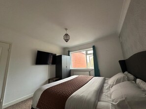 A bedroom with a bed, a flat-screen TV mounted on the wall, a window with curtains, and a ceiling light fixture. - Manchester Retreat With 3 Bedrooms (Manchester)