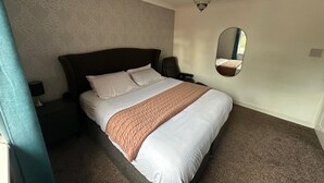 Una cama bien hecha con cabecera, una mesita de noche con lámpara, un espejo en la pared y suelo cubierto de alfombra.