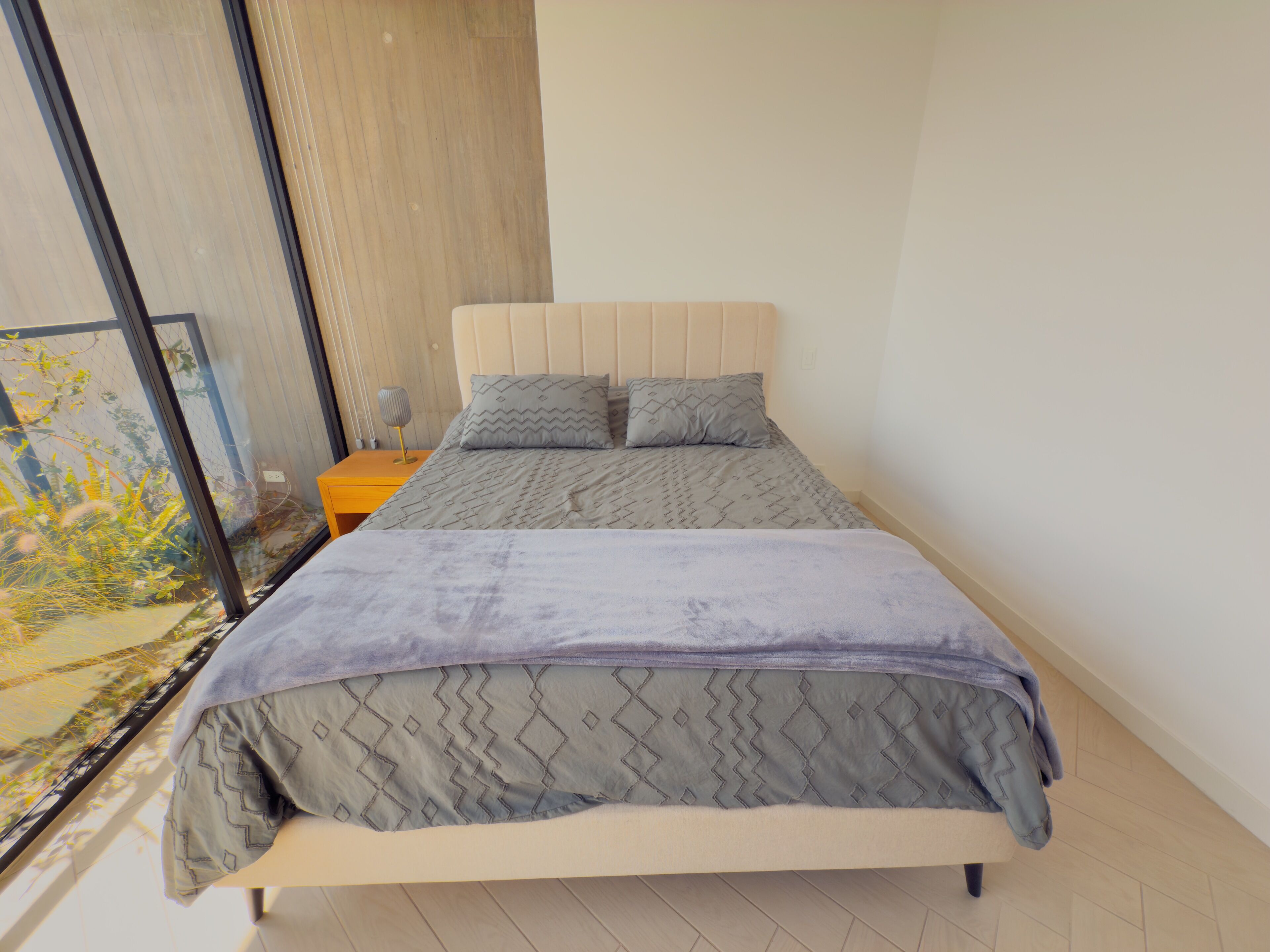Un dormitorio con cama, mesita de noche y ventana con vista a follaje.