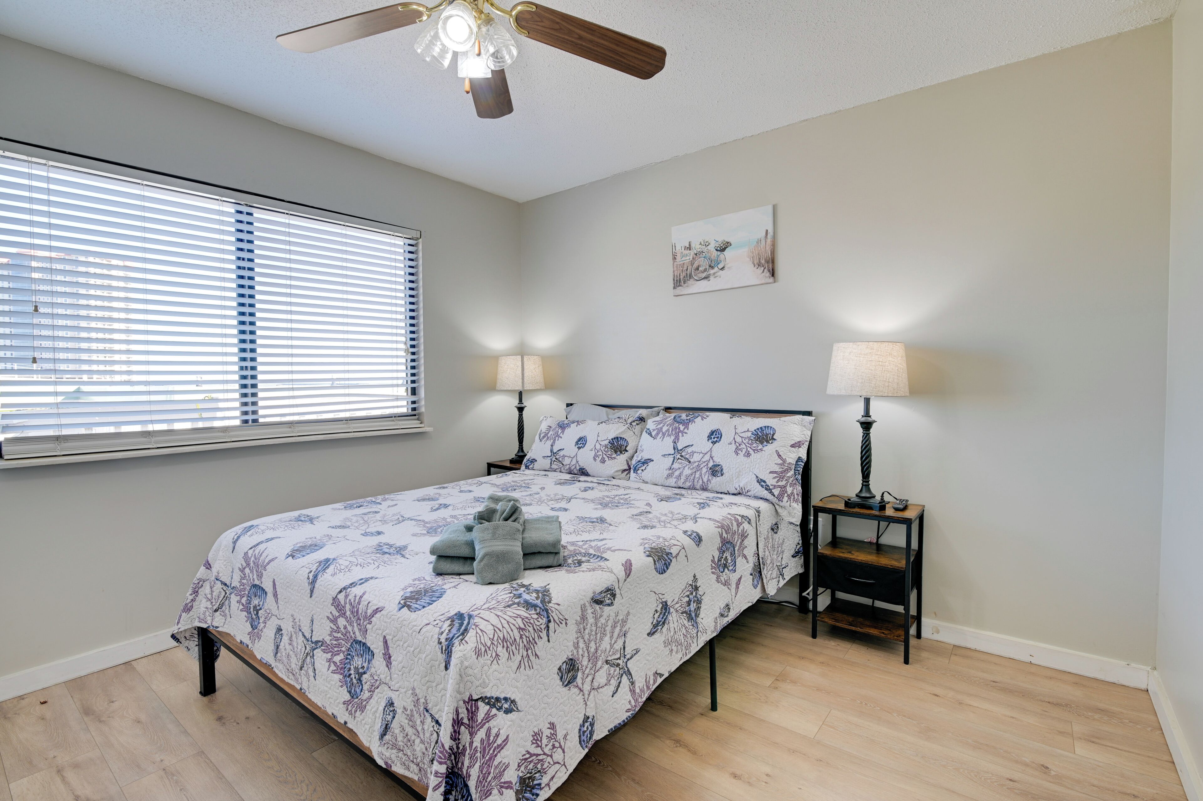 A bedroom with a bed, two bedside tables with lamps, a ceiling fan, and a window with blinds.