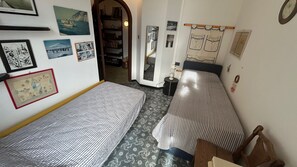 Kapea hotellihuone, jossa on kaksi sänkyä, pieni puinen tuoli, peili ja hylly, jossa on erilaisia esineitä.