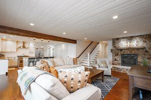 A modern living room with a stone fireplace, wooden ceiling beams, and a kitchen in the background.
