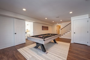 A pool table in a room with wooden flooring, white walls, and a staircase leading to another level.
