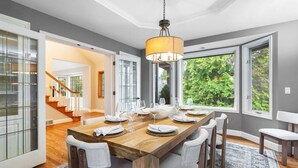 A dining area with a wooden table set for six, surrounded by chairs, with a large window providing a view of greenery outside.