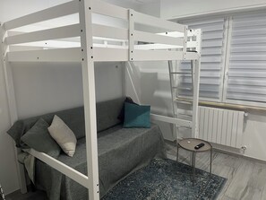 A bunk bed with a grey sofa and pillows, a small round table with a remote, and a radiator.