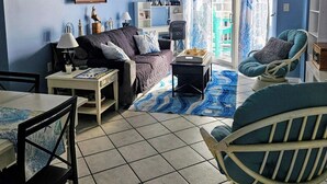 Una sala de estar con sofá, sillón, mesa y escritorio. Hay una ventana con cortinas, una puerta que da al exterior y una pared con un cartel.