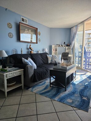 A living room with a sofa, a coffee table, a side table, a lamp, decorative items, and a view of a balcony. - 2-bedroom condo on North Topsail Beach with amazing views of the Atlantic Ocean (North Topsail Beach)