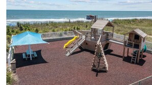 A playground with a slide, climbing structure, and a small wooden house, overlooking a beach.