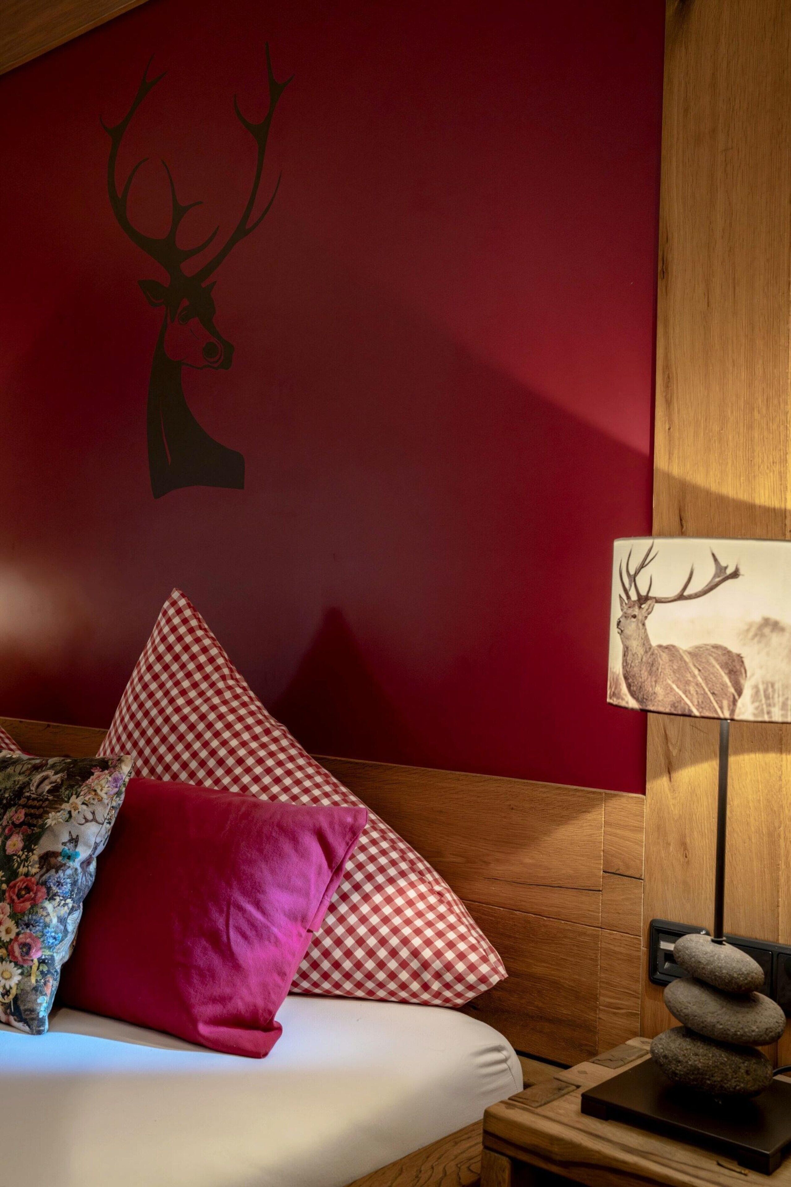 A bedroom with a red wall, a wooden headboard, a bed with white bedding, red and checkered pillows, a decorative deer wall decal, and a lamp with a deer print on the nightstand.