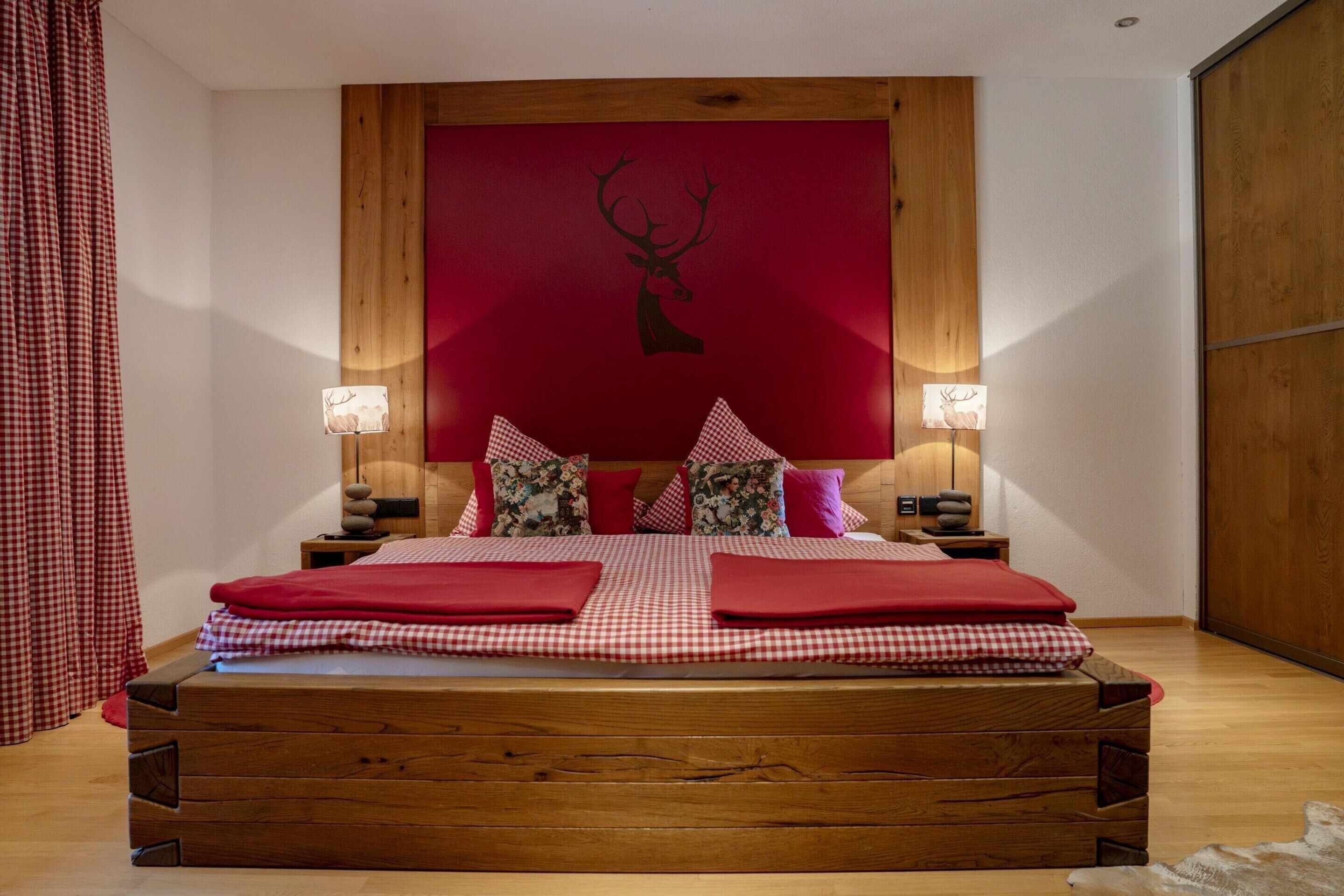 A bedroom with a wooden bed frame, a red wall with a deer head decoration, and two bedside lamps.