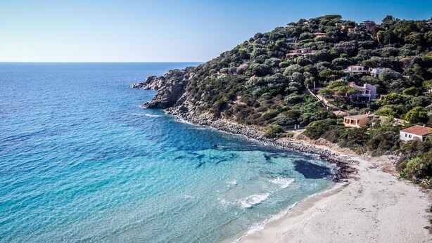 Un paisaje costero con mar azul cristalino, una playa arenosa y una colina con edificaciones.