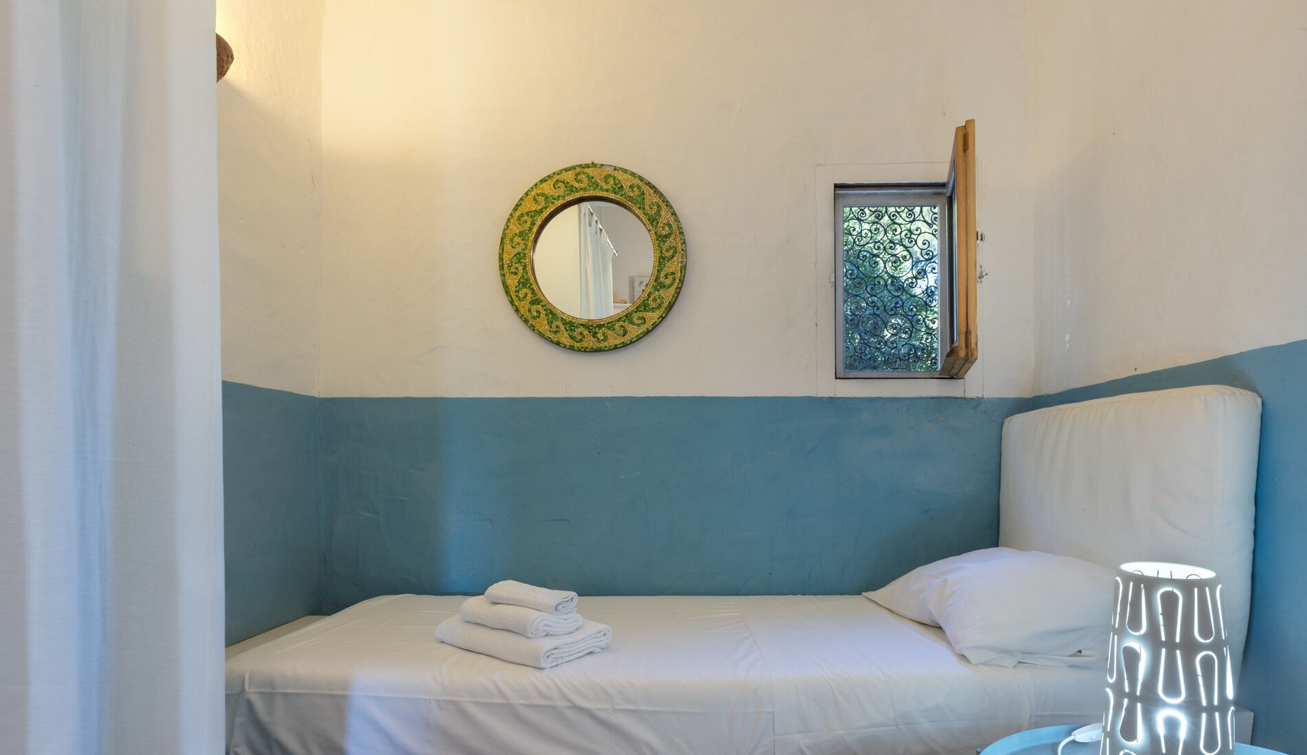 Un dormitorio con una cama, un espejo fijado a la pared, una ventana con barandillas de hierro decorativas y una lámpara de noche.