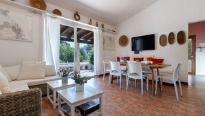 Una sala de estar moderna con sofá, mesa de comedor, sillas y televisión. Cuenta con una ventana con cortinas, un televisor montado en la pared y vistas al exterior.