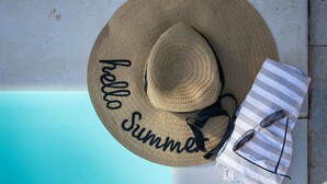 Un sombrero de sol con la leyenda "Hello Summer", gafas de sol y una toalla a rayas sobre una silla de la piscina.