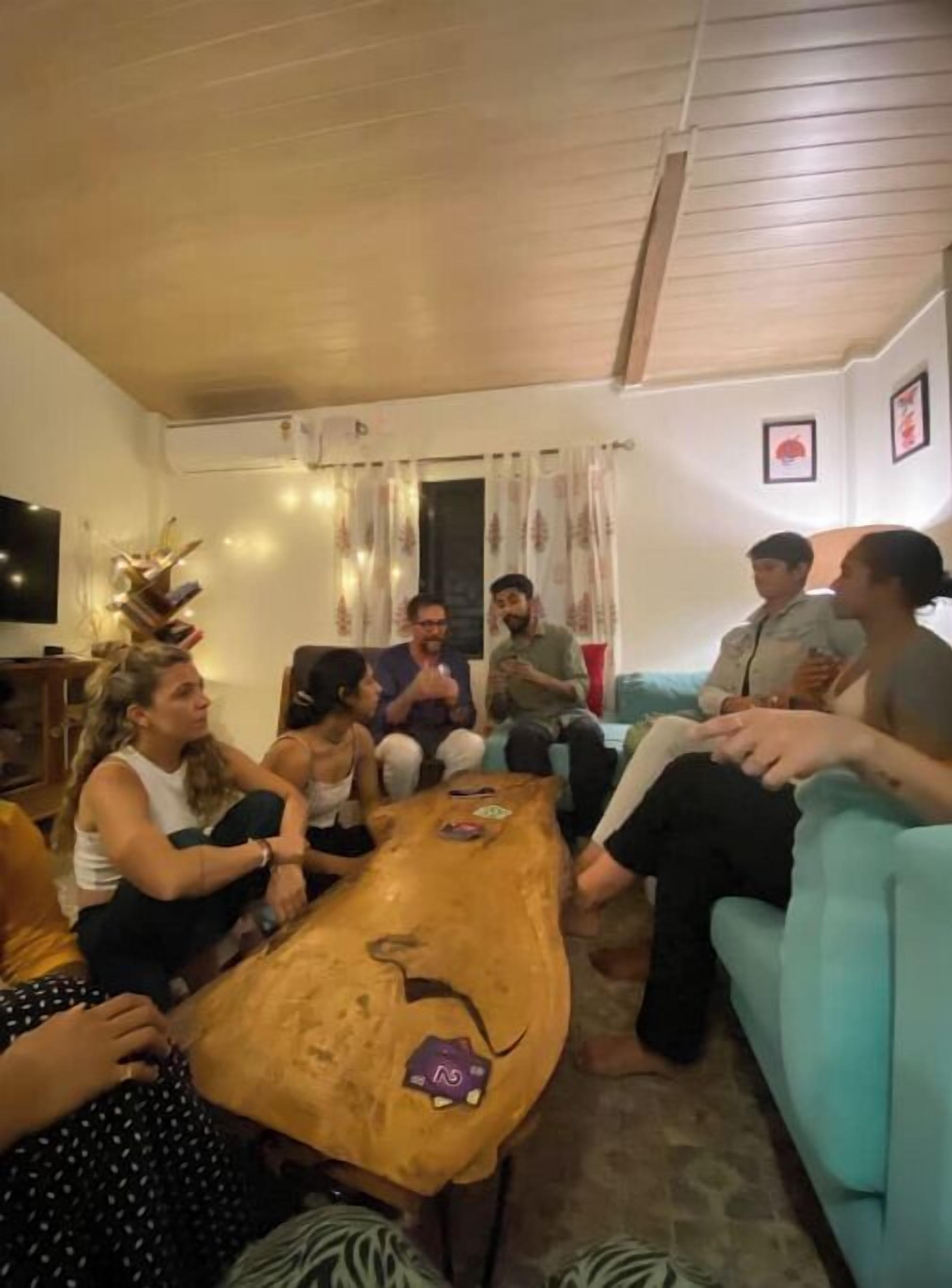 A group of people seated around a wooden coffee table in a living room with a television, curtains, and a light blue couch.