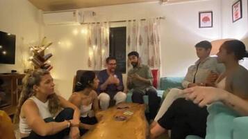 A group of people seated around a wooden coffee table in a living room with a television, curtains, and a light blue couch.