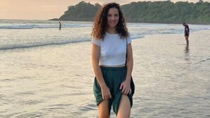 Escena de playa con una persona de pie en el agua, una costa lejana y un cielo nublado.