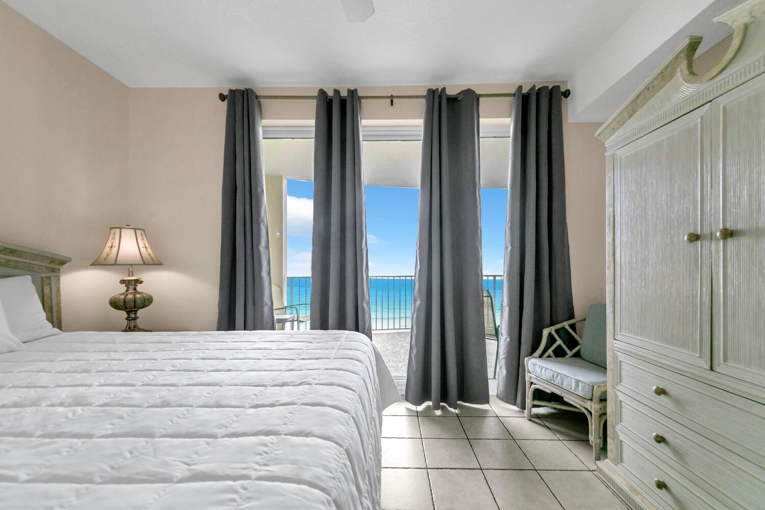 Quarto de hotel com uma cama grande, mesa de cabeceira com abajur, guarda-roupa e varanda com vista para o mar.