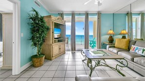 A spacious living room with a view of the ocean, a ceiling fan, a television, and a sofa with a 'HAPPY' pillow.